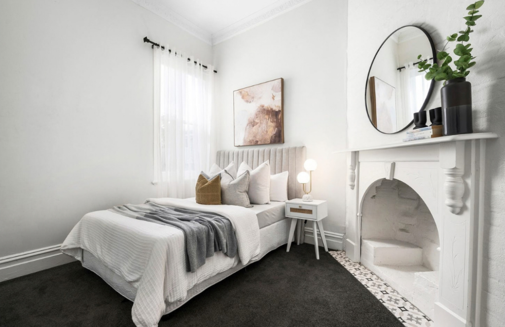 Neutral bedroom with grey upholstered bed, layered cushions, round mirror and original fireplace in a house in Brunswick.