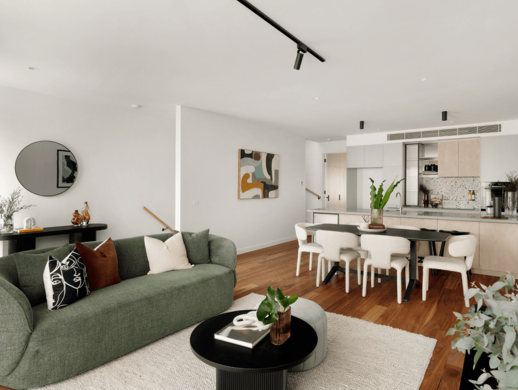 Open-plan living and dining space with curved green sofa, round black coffee table, cream dining chairs and warm timber flooring in the house in Brunswick.
