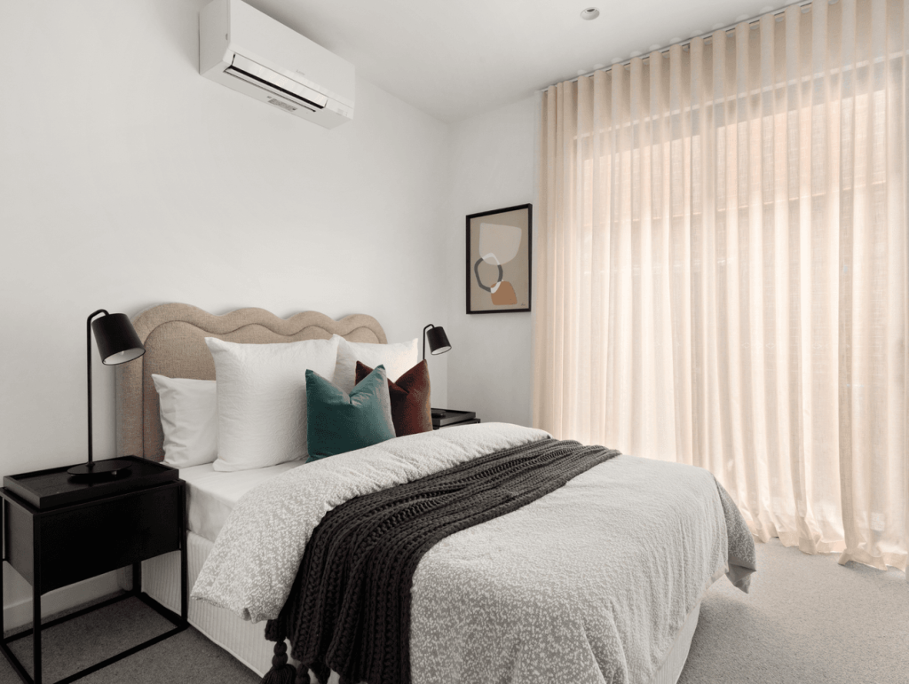 Light-filled bedroom with curved bedhead, white bedding, dark knitted throw and teal and rust cushions in the house in Brunswick.