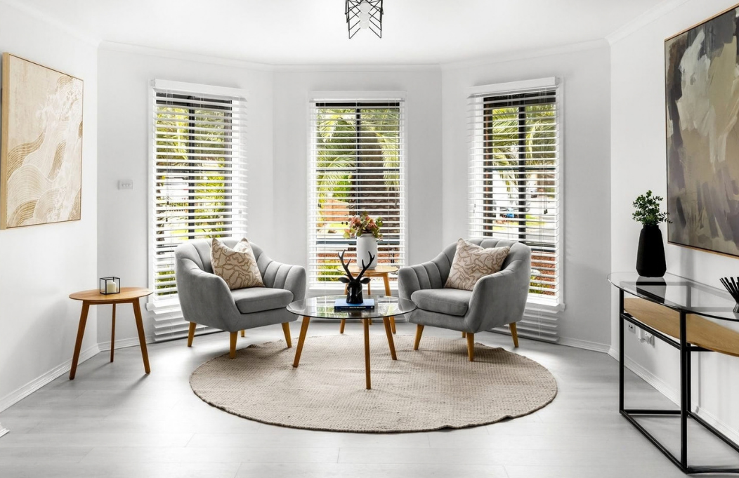 Bright bay-window sitting area styled with two grey armchairs, timber tables and neutral textures.